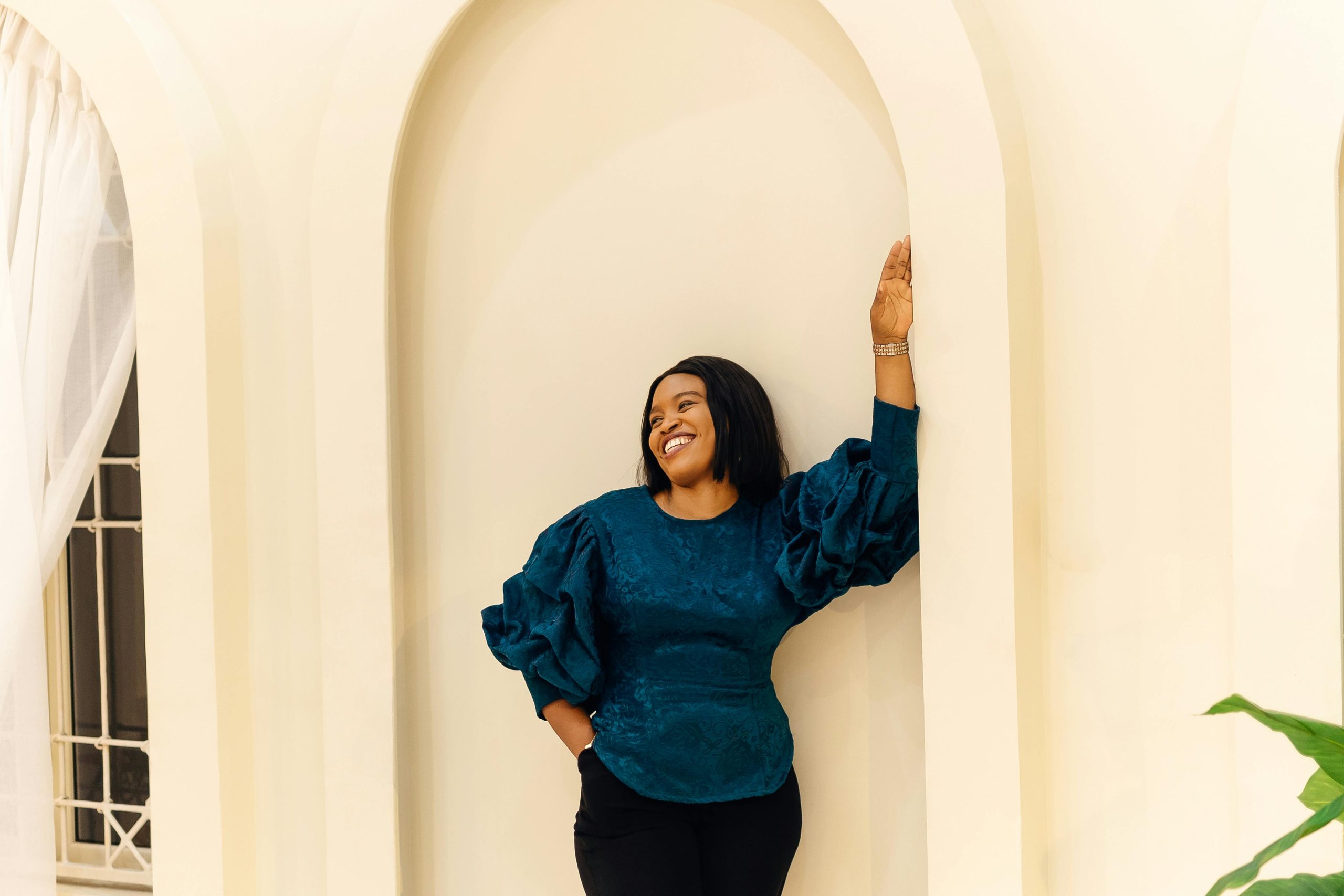 A cheerful woman in Nigeria poses elegantly in a beautiful blue top against a cream wall.